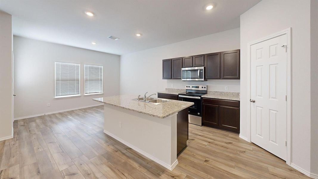 Kitchen featuring stainless steel appliances, dark wood finish cabinets, an island with sink, light stone countertops, and light wood-type flooring
