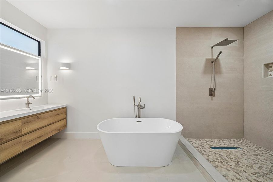 Freestanding soaking tub with floor-mounted faucet, floating wood-finish vanity, backlit mirror, walk-in shower with pebble-stone flooring, and wall-mounted rainfall showerhead Freestanding soaking tub with floor-mounted faucet, floating wood-finish vanity, backlit mirror, walk-in shower with pebble-stone flooring, and wall-mounted rainfall showerhead