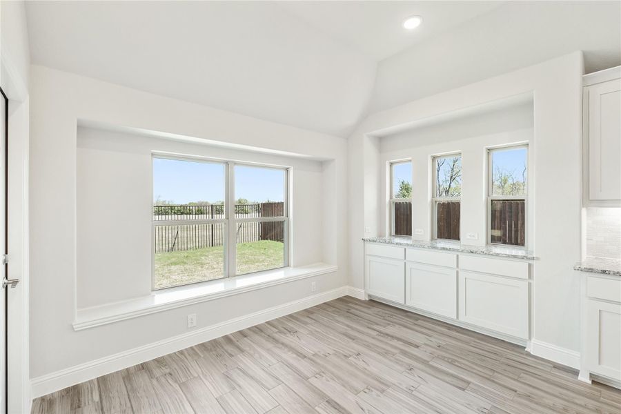 Bright dining area with large windows, white cabinets, granite countertop, and light wood-look floors overlooking a fenced backyard.