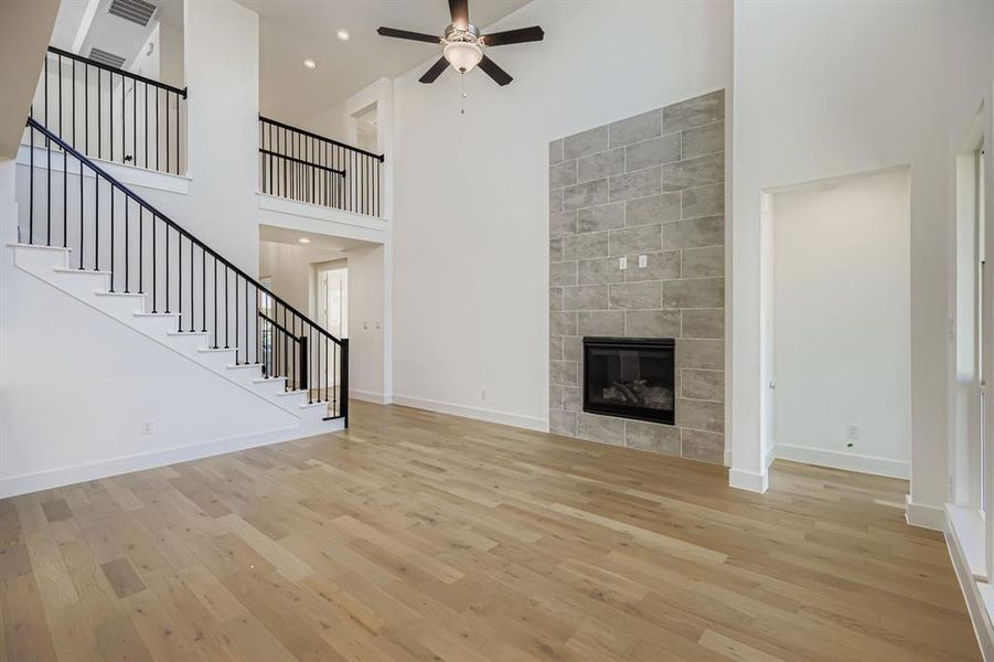 Unfurnished living room with a high ceiling, stairs, light wood-type flooring, a tiled fireplace, and a ceiling fan Unfurnished living room with a high ceiling, stairs, light wood-type flooring, a tiled fireplace, and a ceiling fan