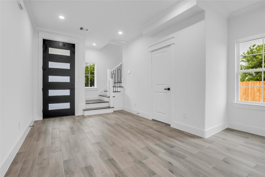 This modern entryway features sleek wood flooring, a stylish black door with frosted glass panels, bright white walls, and ample natural light. A staircase with white risers and dark treads adds a touch of elegance.
