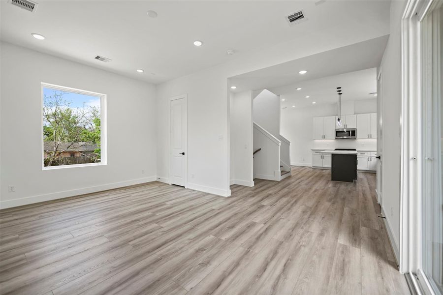 This view from the living area to the kitchen showcases a bright, open-concept living space with modern LVP flooring. The room flows seamlessly into a sleek kitchen featuring white cabinetry, a central island, and stainless steel appliances. Large windows provide natural light, and there's a view of the outdoors.