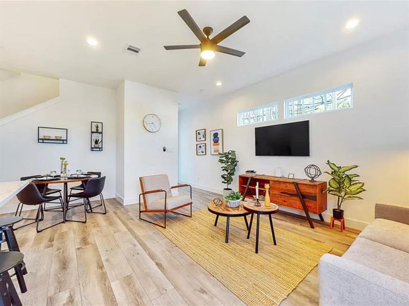 A modern and spacious living area with an open floor plan that seamlessly connects to the kitchen. Natural light floods the room, illuminating the warm wood floors and the contemporary furniture.