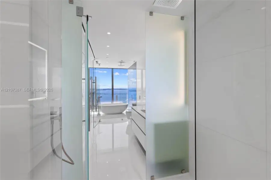 Floor-to-ceiling glass paneling, floating double vanity, freestanding soaking tub, large-format tile flooring, and integrated ceiling rain showerhead