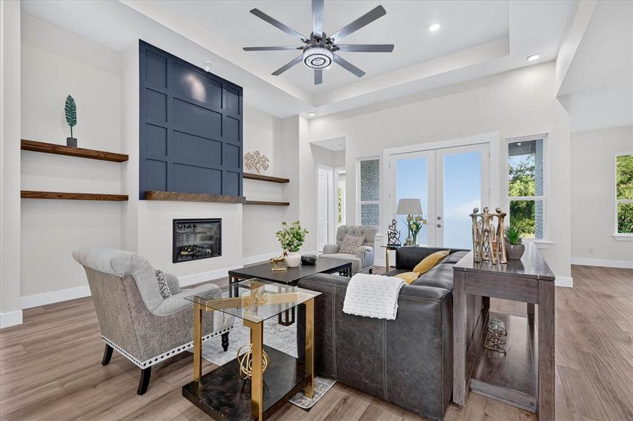 Living area featuring a floor-to-ceiling accent wall with a fireplace, wood-finish flooring, a tray ceiling, and a contemporary ceiling fan Living area featuring a floor-to-ceiling accent wall with a fireplace, wood-finish flooring, a tray ceiling, and a contemporary ceiling fan