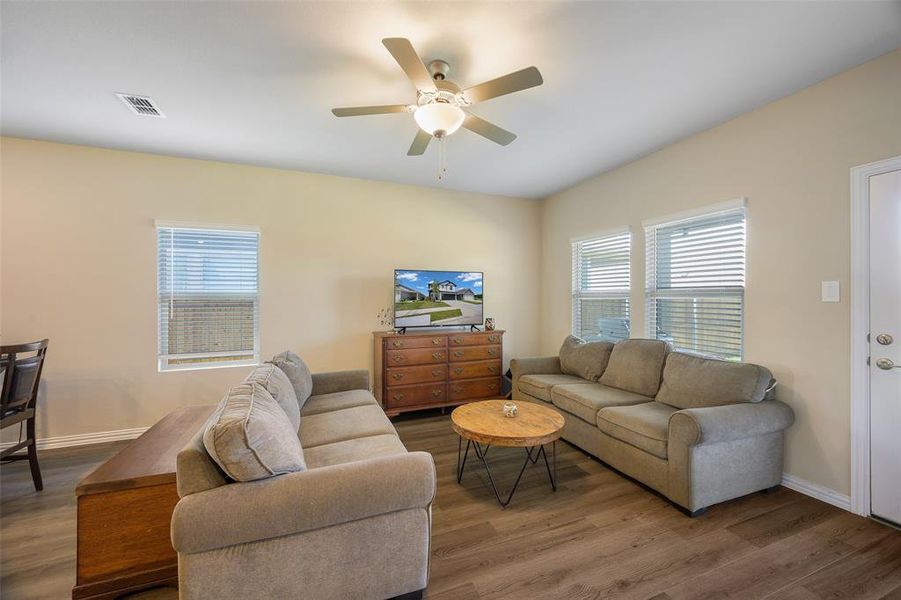 Spacious living area featuring wood-finish flooring, two windows with blinds, and a ceiling fan with integrated lighting Spacious living area featuring wood-finish flooring, two windows with blinds, and a ceiling fan with integrated lighting