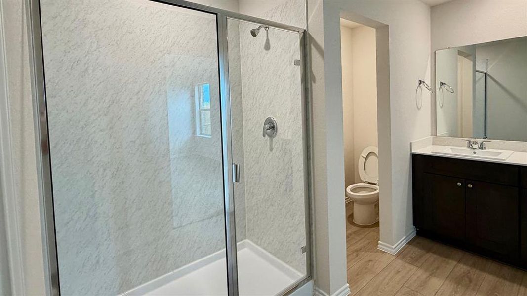Shower enclosure with a clear glass door and a white patterned surround, wood-finish flooring, a dark wood-finish vanity with a light-colored countertop, and a rectangular mirror