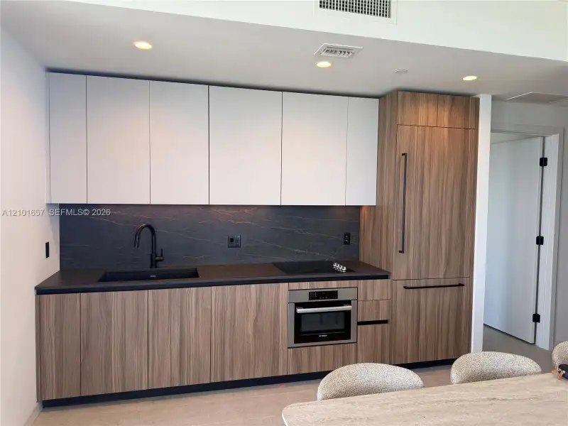 Modern kitchen featuring two-tone cabinetry with wood-finish lower cabinets, matte white upper cabinets, integrated refrigerator, stone backsplash, and recessed lighting