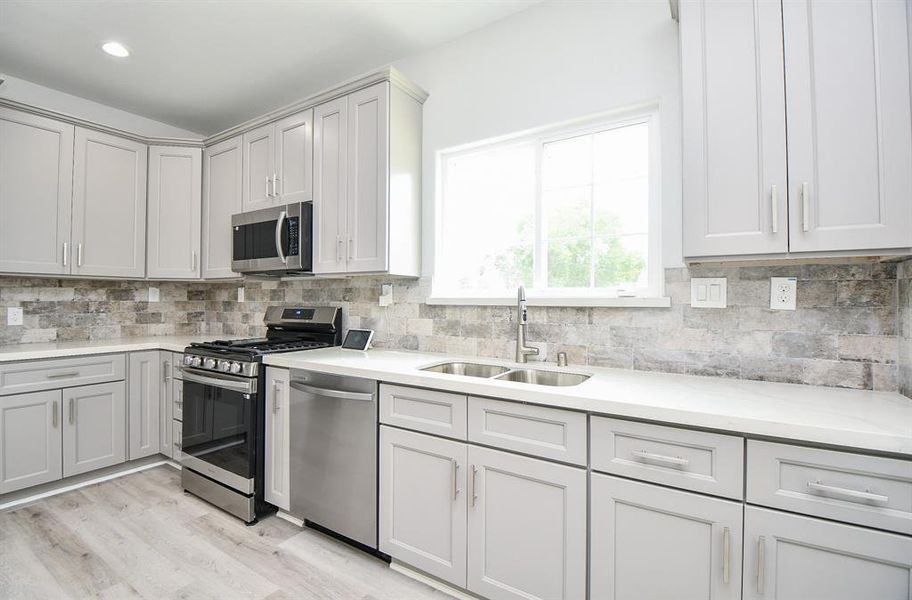 This is a modern kitchen featuring white cabinetry, stainless steel appliances, a gas range, and a backsplash with natural stone tiles. A large window allows for ample natural light.