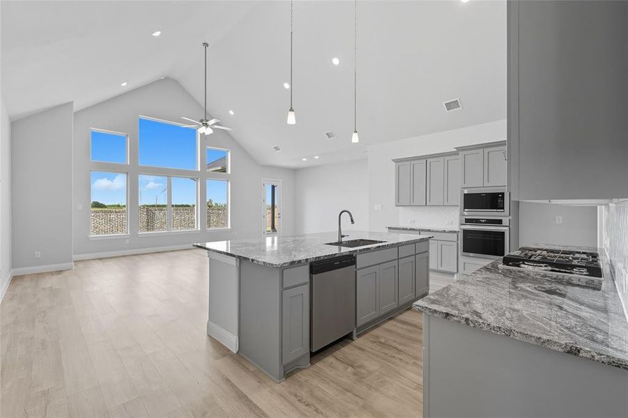 Kitchen featuring high vaulted ceiling, light stone counters, gray cabinetry, an island with sink, and light wood-type flooring Kitchen featuring high vaulted ceiling, light stone counters, gray cabinetry, an island with sink, and light wood-type flooring