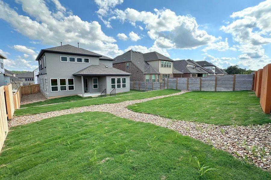 A spacious backyard with pathways of gravel and green grass, bordered by privacy fences and modern homes.