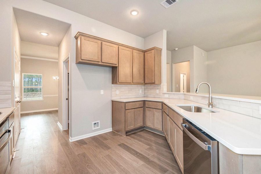 This photo showcases a modern kitchen with natural wood cabinets, a sleek white countertop, and stainless steel appliances. The space features an open layout with a view into an adjacent room, enhancing the flow and natural light.