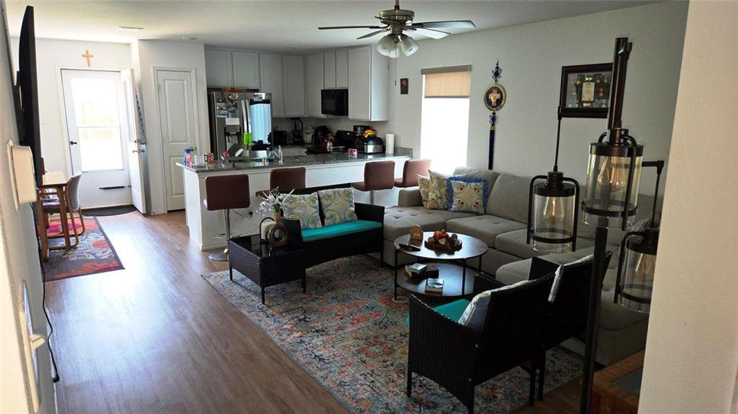 Open-concept living area featuring wood-finish flooring, a kitchen island with seating, upper and lower white cabinetry, a built-in microwave, and a ceiling fan