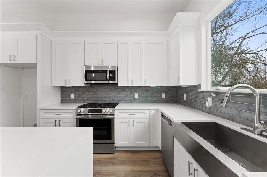 This modern kitchen features sleek white cabinetry, stainless steel appliances, and a large window providing natural light. The gray tile backsplash and spacious countertops offer a stylish and functional cooking space.