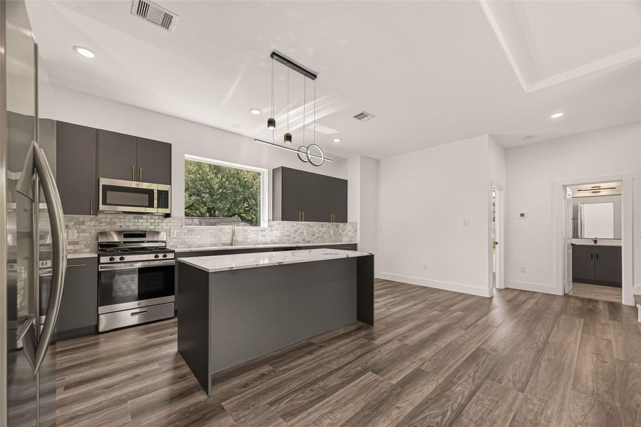 This modern kitchen features sleek dark cabinetry, stainless steel appliances, and a large central island with a marble countertop. The space is brightened by recessed lighting and a large window, with elegant vinyl flooring extending into an open area. This modern kitchen features sleek dark cabinetry, stainless steel appliances, and a large central island with a marble countertop. The space is brightened by recessed lighting and a large window, with elegant vinyl flooring extending into an open area.