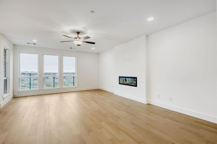 Spacious room featuring light wood-finish flooring, an integrated linear electric fireplace, and three large windows with a metal balcony railing