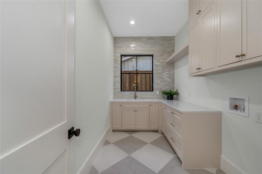 Laundry area featuring checkered flooring, light cabinetry with brass-toned hardware, and a single basin sink with a black-framed window and tile backsplash