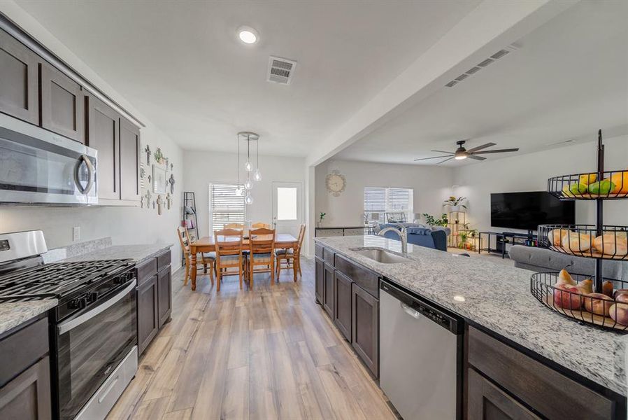 Kitchen with dark brown cabinetry, stainless steel appliances, light stone counters, pendant lighting, and light wood finished floors