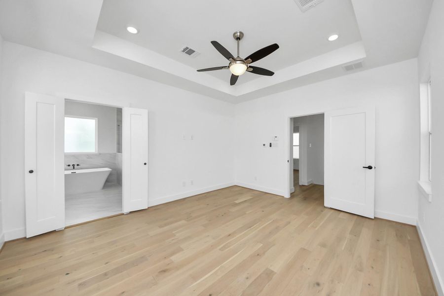 This room features a spacious, bright design with light wood flooring and white walls. It includes a ceiling fan and recessed lighting. The room opens to a modern bathroom with a freestanding tub, visible through a set of double doors.