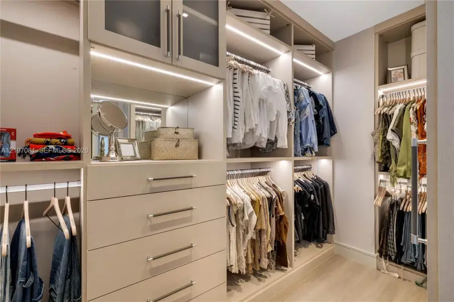 Custom-built walk-in closet featuring floor-to-ceiling cabinetry, integrated LED lighting, glass-front upper storage, soft-close drawers with metallic pulls, and wood-finish flooring Custom-built walk-in closet featuring floor-to-ceiling cabinetry, integrated LED lighting, glass-front upper storage, soft-close drawers with metallic pulls, and wood-finish flooring