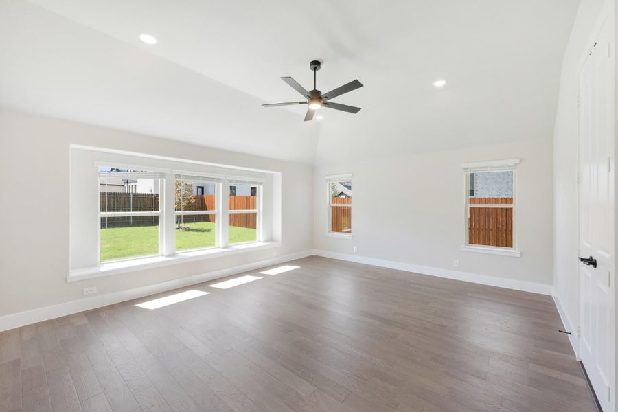 Empty living room with hardwood floors, ceiling fan, vaulted ceiling, and large windows overlooking backyard