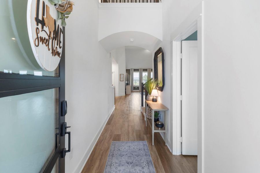 Bright, welcoming entryway with light vinyl plank flooring, high ceilings, and neutral paint. The hallway leads to a spacious living area filled with natural light from large windows. To the right is the downstairs secondary bedroom with full bathroom. Perfect for guests.