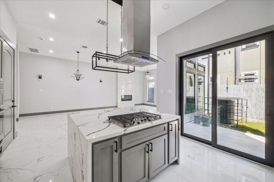 At the heart of the kitchen, the island stands out with a striking waterfall-edge stone surface that serves as both prep and serving space. A gas cooktop paired with a sleek vent hood blends function and style, while pendant lighting adds a modern touch.