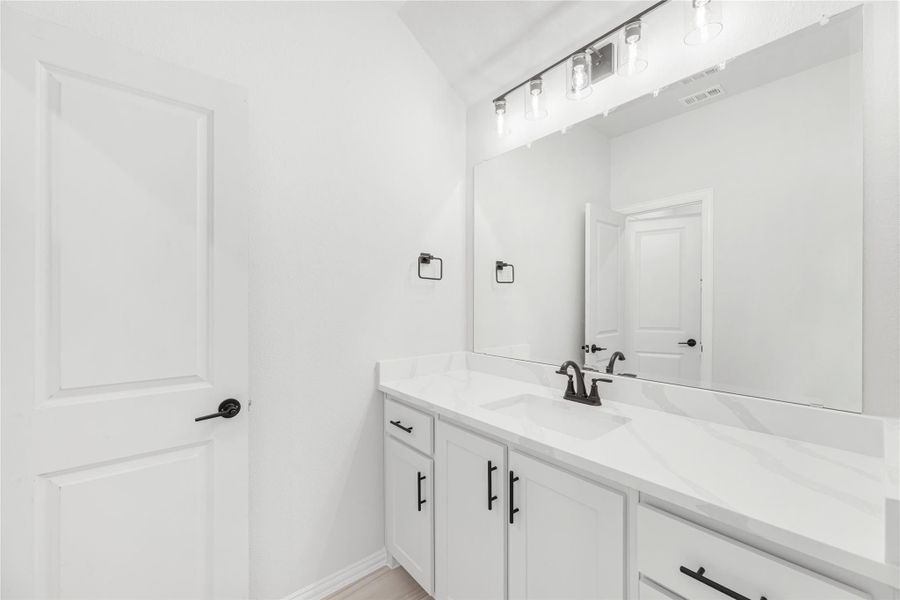 Bathroom vanity with white cabinets, quartz countertop, single sink, black hardware, and large mirror with overhead lighting. Bathroom vanity with white cabinets, quartz countertop, single sink, black hardware, and large mirror with overhead lighting.