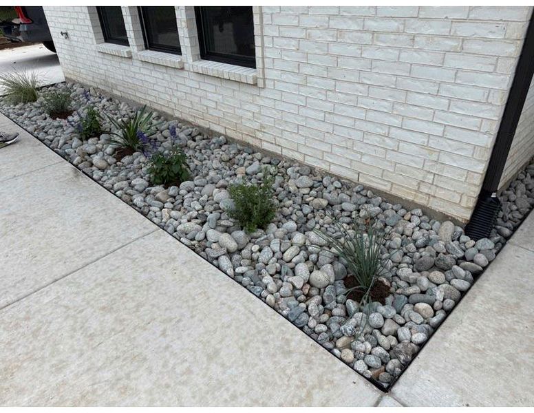 White brick exterior featuring a rock bed with varied plantings, black framed windows, and a concrete pathway