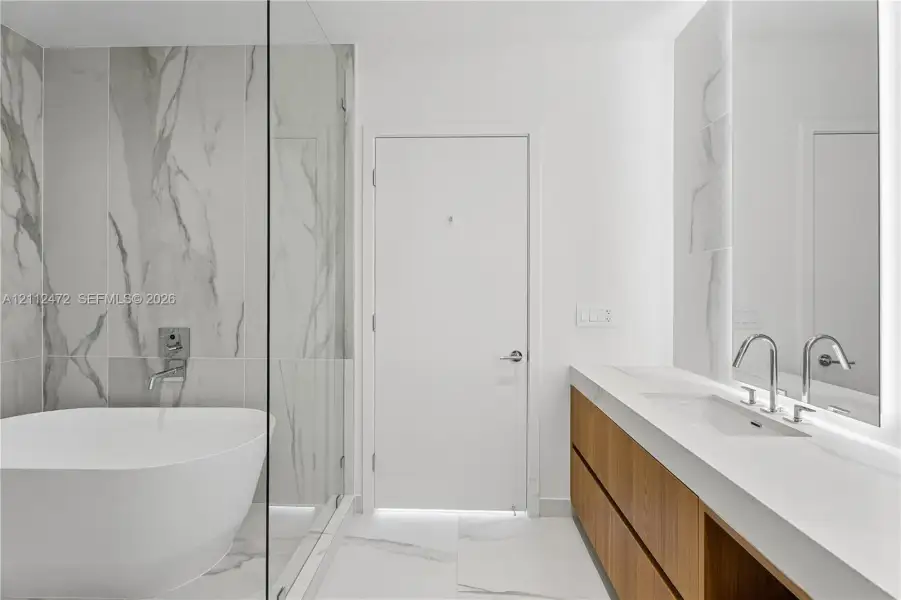 Freestanding soaking tub, walk-in shower with glass enclosure, wall-mounted faucet, dual-sink vanity with wood-finish cabinetry, and oversized stone-patterned wall and floor tiling