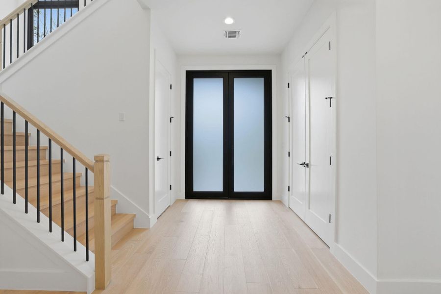 Entry foyer featuring light wood-finish flooring, a modern staircase with natural wood handrails and black metal balusters, and a contemporary black double door with frosted glass panels