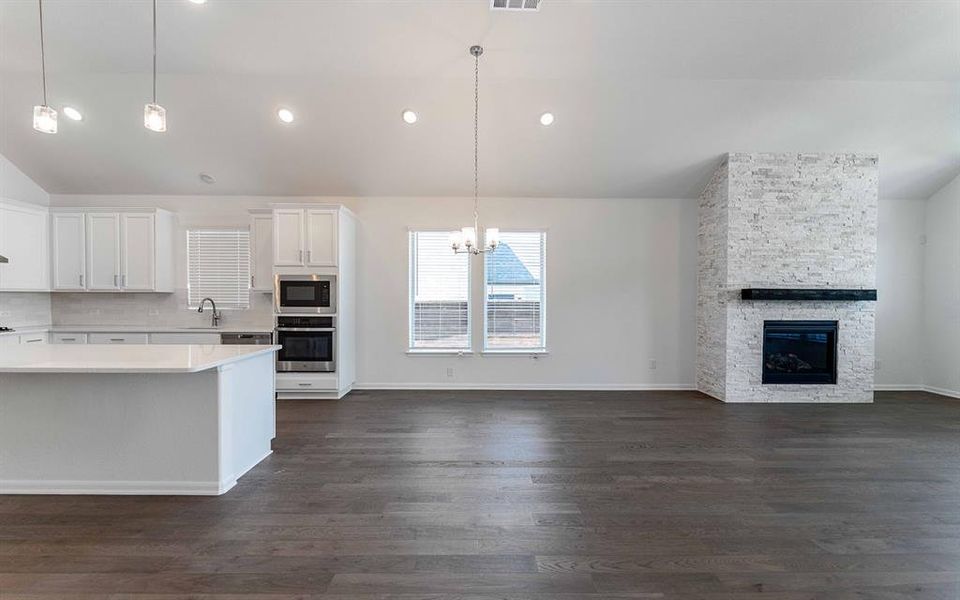 Open-concept living space featuring wood-finish flooring, a contemporary fireplace with stacked stone surround and dark mantel, and a kitchen with white cabinetry, built-in appliances, and a central island