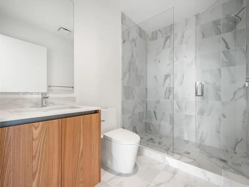 Bathroom featuring a wood-finish vanity with a stone-surfaced countertop, large-format tile flooring, walk-in glass-enclosed shower with floor-to-ceiling tile surround, and a wall-mounted mirror Bathroom featuring a wood-finish vanity with a stone-surfaced countertop, large-format tile flooring, walk-in glass-enclosed shower with floor-to-ceiling tile surround, and a wall-mounted mirror