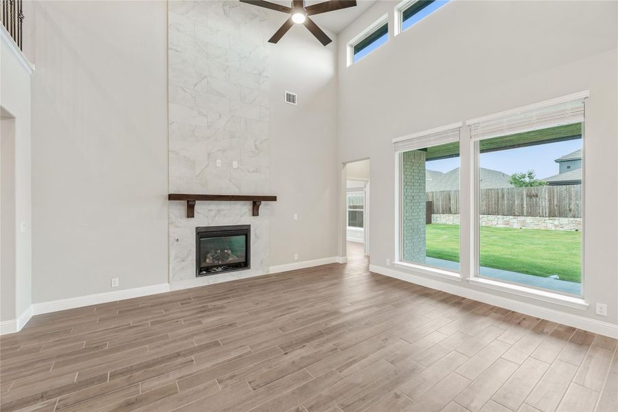 Open living room with two-story ceiling, marble tile fireplace, wood floors, and large windows overlooking backyard