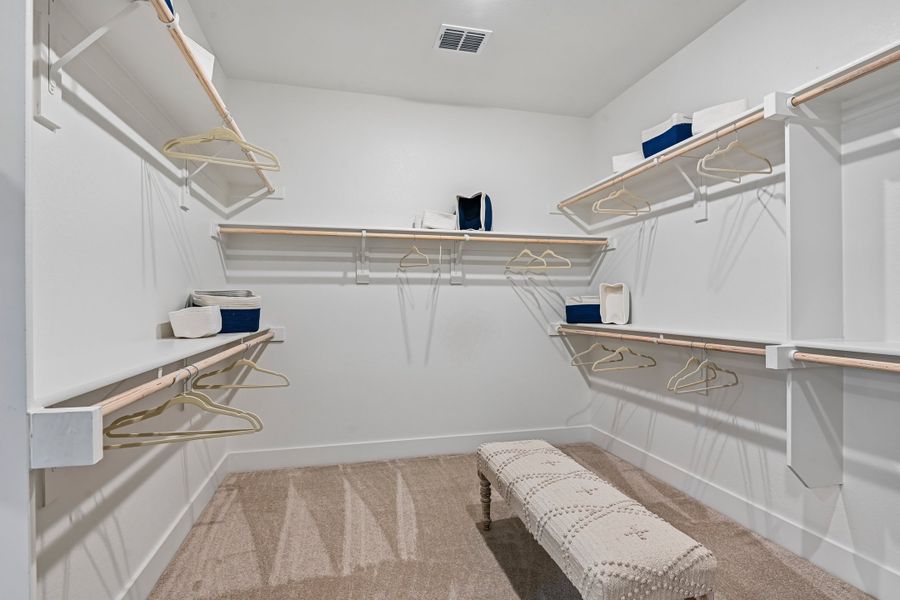 Walk-in closet with white shelving, multiple hanging rods, carpet flooring, and a small upholstered bench in the center.