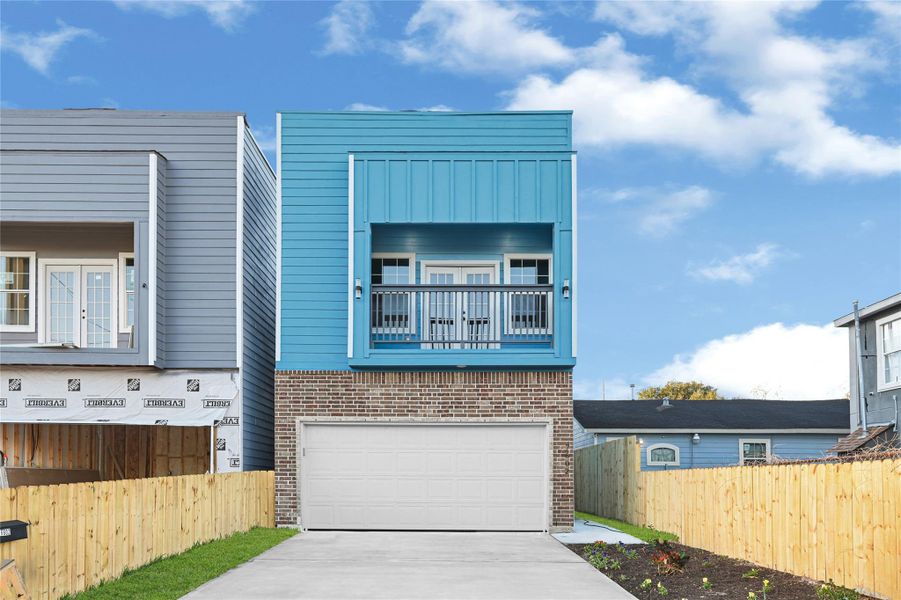 Modern two-story home with a vibrant blue and brick facade. Features a balcony above a spacious garage, and a fenced driveway with a small garden area. Ideal for contemporary living.