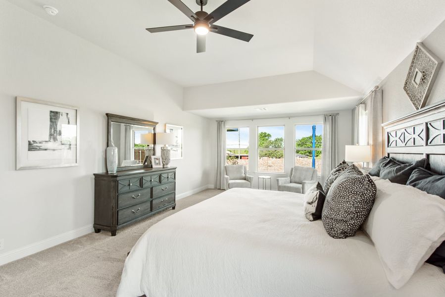 Spacious master bedroom with vaulted ceiling, ceiling fan, dark wood dresser, and large windows with seating area