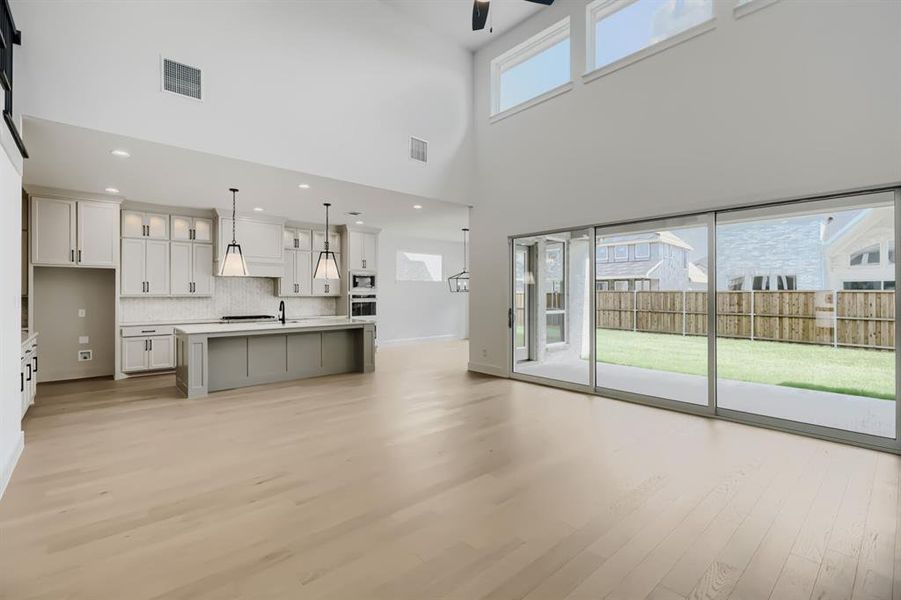 Open-concept living area featuring a two-story ceiling, large sliding glass doors, light wood-finish flooring, and a modern kitchen Open-concept living area featuring a two-story ceiling, large sliding glass doors, light wood-finish flooring, and a modern kitchen