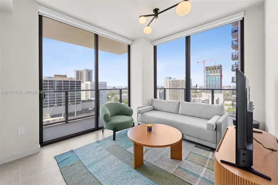 Floor-to-ceiling windows, sliding glass door with access to a private balcony, neutral large-format tile flooring, and a modern multi-bulb chandelier