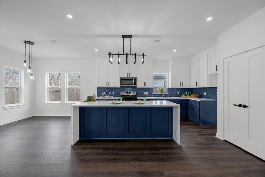 Stylish dark wood floors and bright white walls. It includes a large island with blue cabinetry, matching lower cabinets, and a stylish light fixture. The kitchen is well-lit with natural light from multiple windows, creating a welcoming atmosphere.