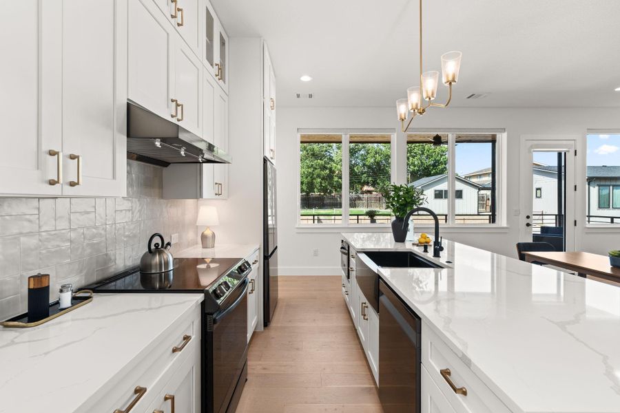 Bright, modern kitchen with white cabinetry, sleek countertops, and a large island featuring a black sink. Stainless steel appliances and stylish pendant lighting enhance the space. Ample natural light streams in through large windows, offering a beautiful view.