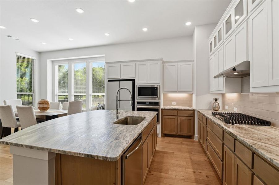 Open-concept kitchen featuring an oversized island with a built-in sink, stainless steel appliances, light wood-finish flooring, white upper cabinetry with glass inserts, and contrasting light wood-finish lower cabinetry Open-concept kitchen featuring an oversized island with a built-in sink, stainless steel appliances, light wood-finish flooring, white upper cabinetry with glass inserts, and contrasting light wood-finish lower cabinetry