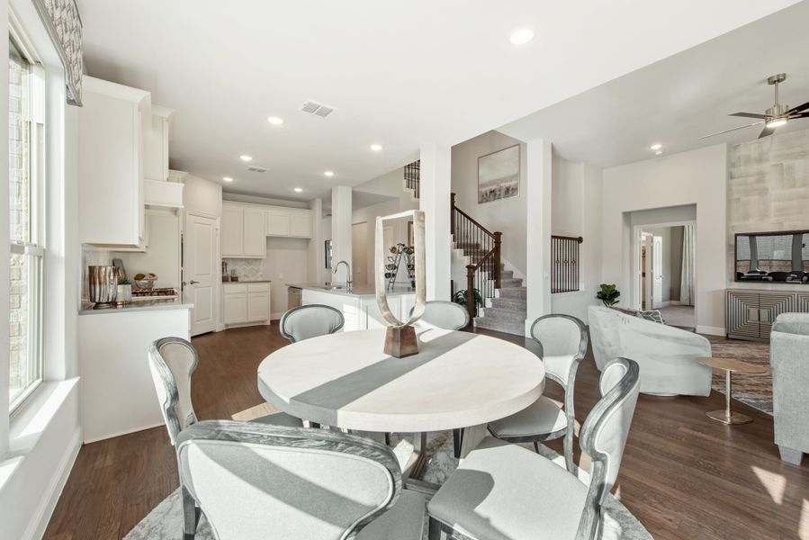Open-plan dining area with round white table, gray chairs, hardwood floors, and kitchen and staircase in background