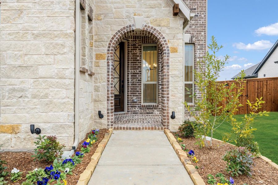 Home entrance with brick arched doorway, stone exterior, concrete walkway, and flower bed landscaping Home entrance with brick arched doorway, stone exterior, concrete walkway, and flower bed landscaping