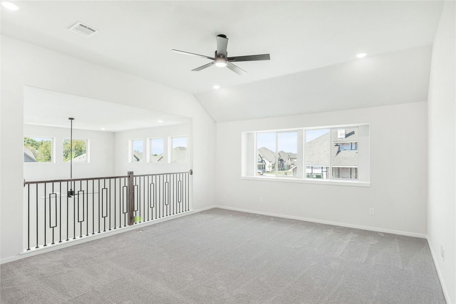 Open upstairs bonus room with vaulted ceiling, ceiling fan, gray carpet, and iron stair railing overlooking lower level Open upstairs bonus room with vaulted ceiling, ceiling fan, gray carpet, and iron stair railing overlooking lower level