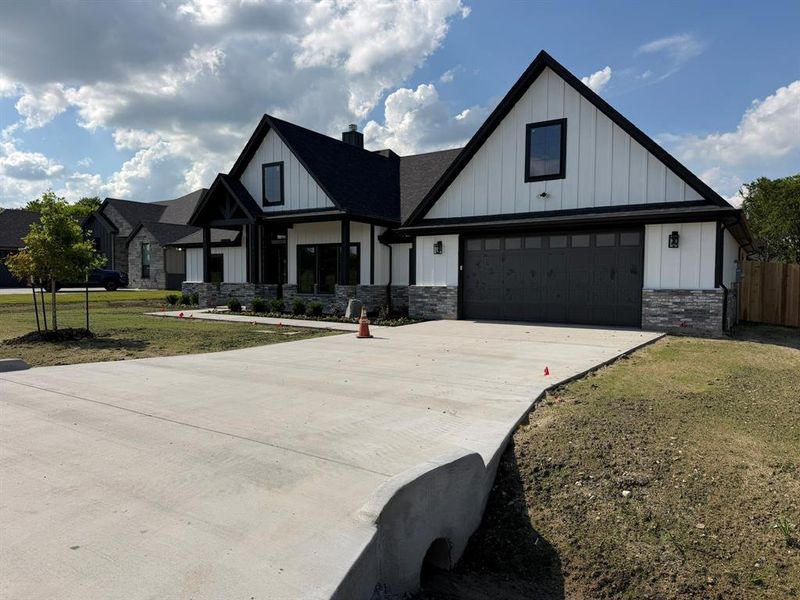 Modern farmhouse exterior featuring white board and batten siding, dark trim, a two-car garage with black door, stone wainscoting, and a covered entry porch with dark wood accents