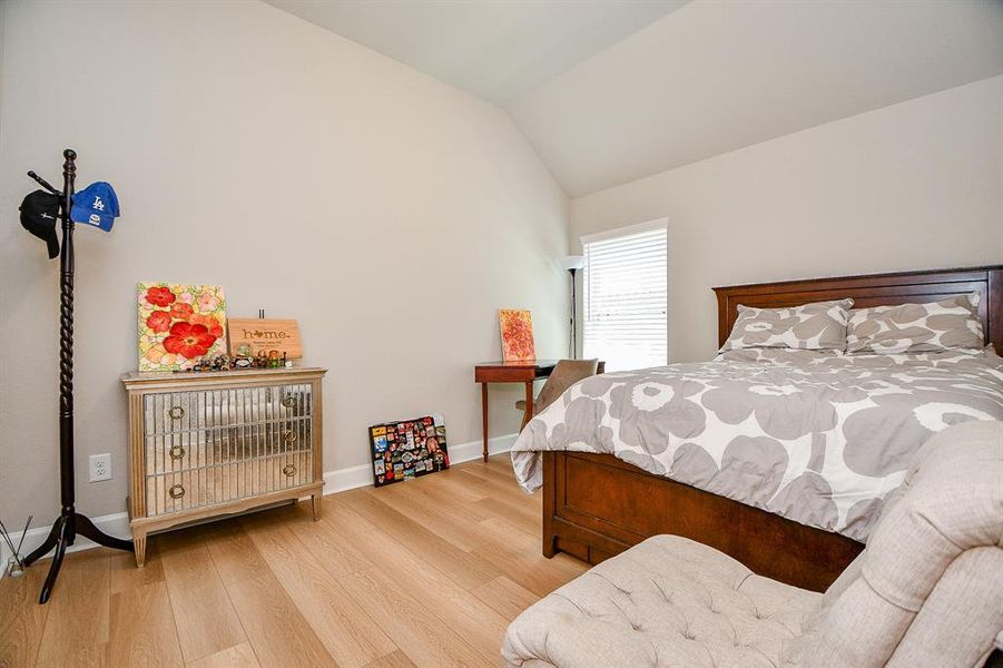 This bedroom features a cozy setup with a wooden bed, floral bedding, and ample natural light from a window. It includes a mirrored cabinet and a small desk, complemented by light hardwood flooring and neutral walls.