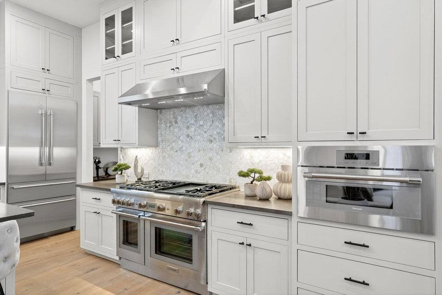 Kitchen featuring high end appliances, glass insert cabinets, white cabinets, under cabinet range hood, and backsplash
