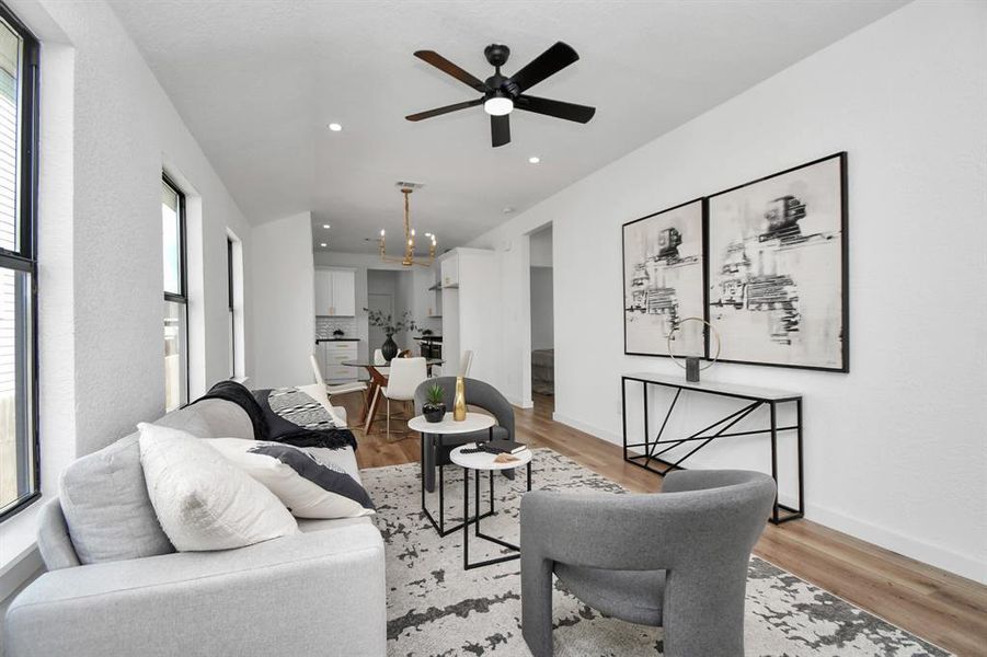 This is a bright, modern dining space featuring a glass table with white chairs and a chic gold chandelier. The room opens to a sleek, white kitchen with stainless steel appliances. Large windows provide natural light, and light hardwood floors run throughout.