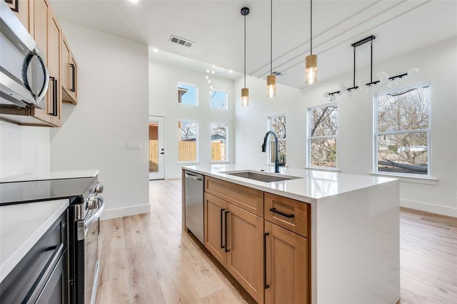 Kitchen featuring brown cabinetry, stainless steel appliances, light wood-type flooring, decorative light fixtures, and light stone countertops